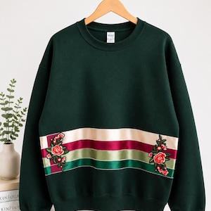 May include: A dark forest green crewneck sweatshirt featuring a horizontal band of layered satin ribbons in cream, burgundy, and sage green. Embroidered floral motifs with red roses and green leaves are positioned on the left and right sides.
