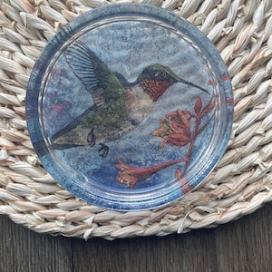 Op de afbeelding: Een ronde glazen onderzetter met een afbeelding van een kolibrie in vlucht, tegen een blauw-grijze achtergrond. De kolibrie heeft groene, rode en zwarte details. De onderzetter ligt op een geweven, lichtbruine mat.