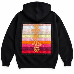 Può includere: Una felpa nera con cappuccio caratterizzata da una grande toppa quadrata sul retro. La toppa è composta da strisce orizzontali satinate nei toni del bianco, beige, giallo, rosa, arancione e rosso, con un disegno floreale arancione.