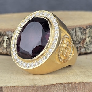 May include: A gold-colored ring with a large, oval, dark purple gemstone. The gemstone is surrounded by a halo of small, clear stones. The ring has a detailed design on the side and is set on a wooden surface.