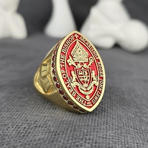 Könnte beinhalten: Ein goldfarbener Ring mit einem roten ovalen Mittelstück. Das Mittelstück zeigt ein Wappen mit den Worten "The Seal of the Bishop" und "Archbishop Roger John Lewis". Der Ring ist mit kleinen roten Edelsteinen besetzt.