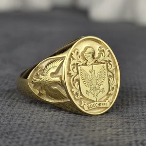 May include: A gold signet ring with an oval face featuring a detailed coat of arms design. The ring has a polished finish and a wide band. The word "Bocchieri" is inscribed at the bottom of the face.