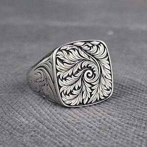 Puede incluir: Un anillo de sello de plata con un intrincado diseño floral grabado. La cara cuadrada del anillo presenta un diseño detallado de hojas y vides arremolinadas. La banda del anillo también tiene detalles grabados.