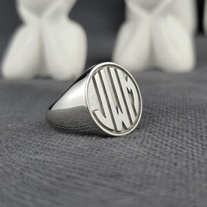 May include: A polished silver signet ring with a circular face engraved with the initials "JWM" in a vertical striped pattern. The ring is set against a grey background with blurred white sculptures.
