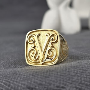 Könnte beinhalten: Ein goldfarbener Siegelring mit dem Buchstaben "V" in einer verzierten, stilisierten Schriftart. Der Ring hat eine quadratische Oberfläche mit dekorativen Verzierungen und einem detaillierten Band. Der Ring ist wahrscheinlich aus Gold oder einem goldfarbenen Metall.