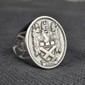 May include: A silver signet ring with an oval face. The face features a detailed family crest with the name "Johnston" inscribed on a banner. The ring has a polished finish and a wide band.