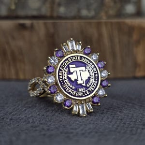 Könnte beinhalten: Ein goldfarbener Ring mit dem kreisförmigen Emblem der Tarleton State University in Stephenville, Texas, und der Jahreszahl 1899. Die Mitte ist von einem Kranz aus abwechselnd runden violetten und klaren Steinen sowie klaren Baguette-Steinen umgeben.