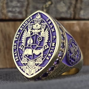 Könnte beinhalten: Ein goldfarbener Ring mit einer großen, schildförmigen Vorderseite und einer violetten Emaille-Einlage. Die Vorderseite zeigt ein Wappen mit Mitra, Krummstab, offenem Buch, Herz und Händedruck, umgeben von dem Text SEAL OF THE BISHOP SEAN T. MILES.