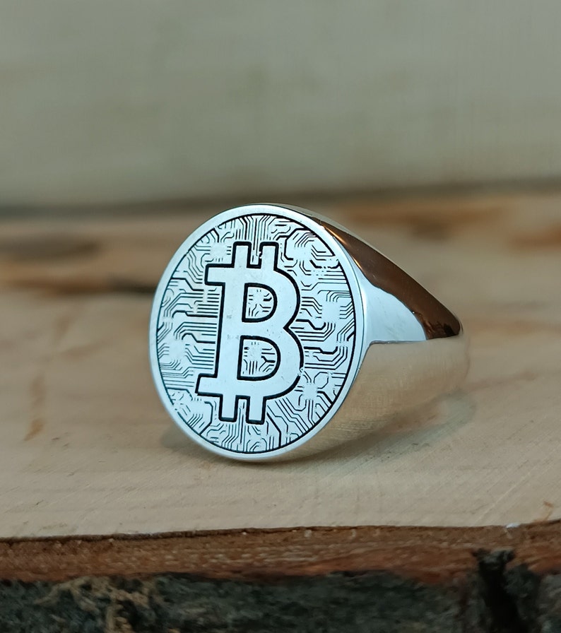 May include: A silver ring with a circular design featuring a raised Bitcoin symbol on a textured background.