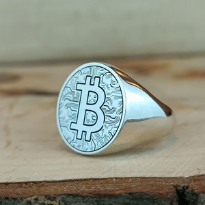 May include: A silver ring with a circular design featuring a Bitcoin symbol and a circuit board pattern.