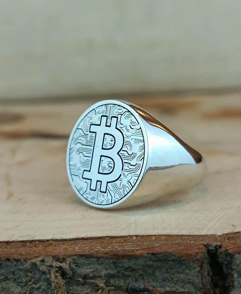 May include: A silver ring with a circular design featuring a Bitcoin symbol and a circuit board pattern.