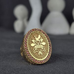 May include: A gold-colored ring with an oval face, featuring a detailed emblem with text and symbols. The ring is adorned with small, red gemstones around the face. The text includes "BISHOP EMERITUS 1957 AD" and "CHIEF APOSTLE HUE L. ROGERS PRESIDING".