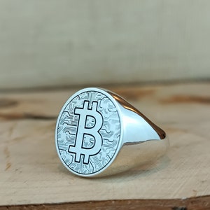May include: A silver ring with a large, round face featuring a raised, detailed Bitcoin symbol. The ring is on a wooden surface.