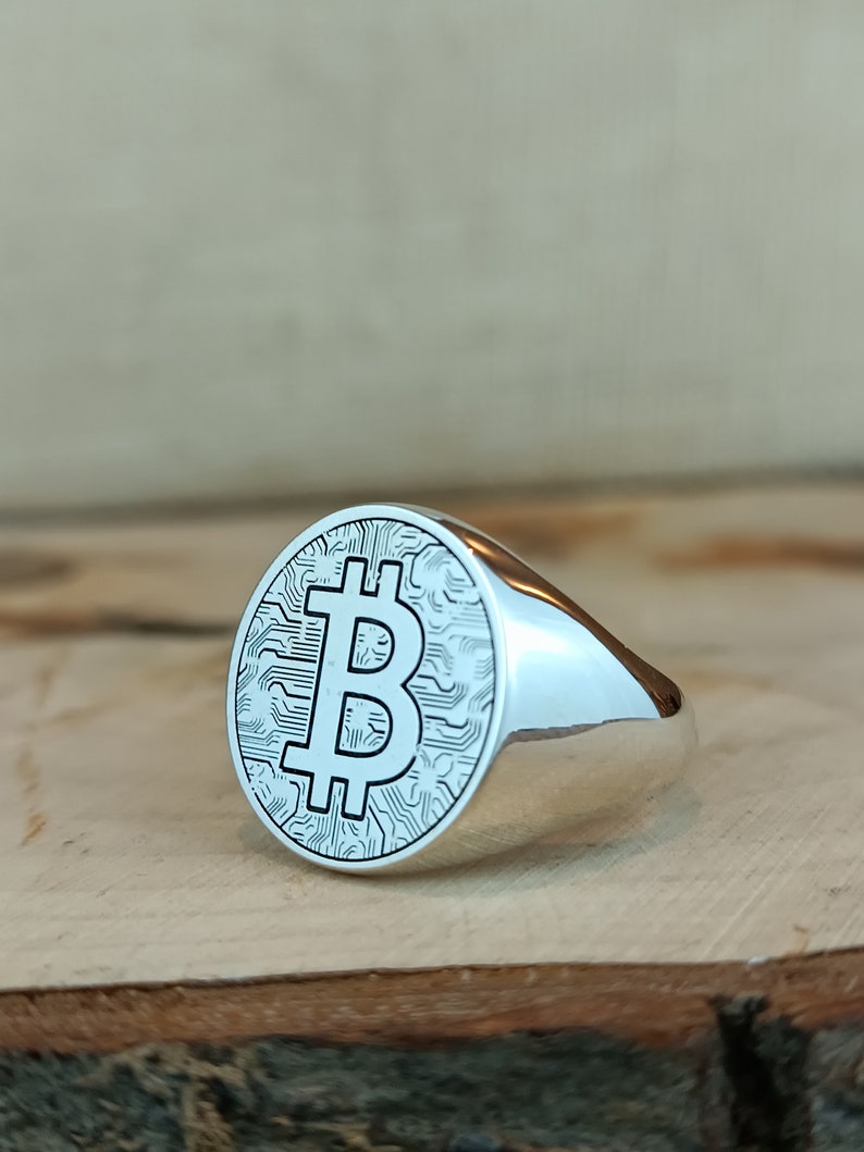 May include: A silver ring with a large, round face featuring a raised, detailed Bitcoin symbol. The ring is on a wooden surface.