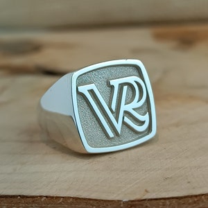 May include: A silver signet ring with a square face featuring the initials "VR" engraved in a bold, stylized font.