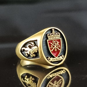 May include: Gold-toned ring with a black and red crest design. The crest features a crown, cross, and the text "Ave Maria". A lion emblem is on the side. The ring is highly reflective.