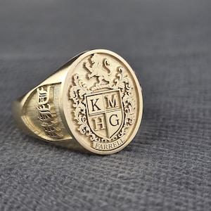 Op de afbeelding: Een gouden zegelring met een ovale voorkant. De voorkant heeft een wapen met een leeuw, de initialen "KM HG" en de naam "Farrell". De ring heeft een gedetailleerd ontwerp en een gepolijste afwerking.