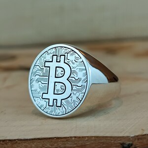 May include: A silver ring with a circular design featuring a Bitcoin symbol on a wood surface.