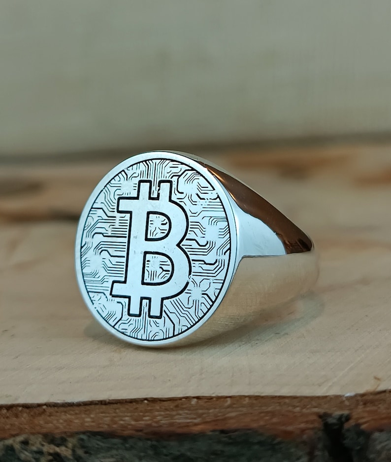 May include: A silver ring with a circular design featuring a Bitcoin symbol on a wood surface.