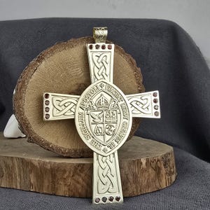 Puede incluir: Un colgante de cruz metálico con intrincados patrones de nudos celtas en los brazos y un emblema circular central. El emblema muestra un escudo con símbolos religiosos y el texto THE SIGILLUM APOSTOLI VINCENT LEROY GREAVES SR.