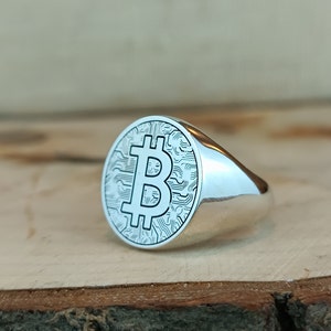 May include: A silver ring with a bitcoin symbol engraved on the face. The ring is oval shaped and has a smooth, polished finish.
