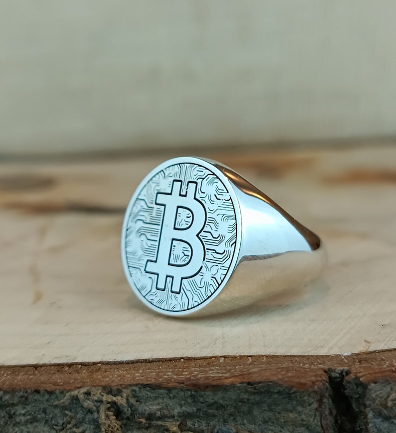 May include: A silver ring with a bitcoin symbol engraved on the face. The ring is oval shaped and has a smooth, polished finish.