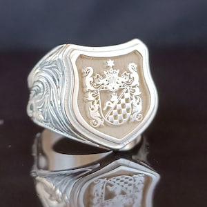 May include: A silver ring with a detailed design featuring a crest with a checkered pattern and a small animal figure. The crest is surrounded by a floral design.