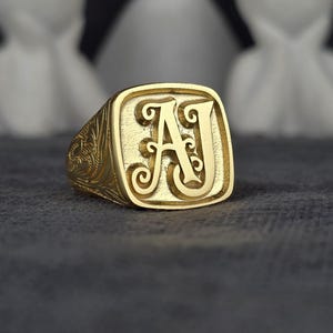 May include: A gold-colored signet ring with the initials "AJ" in a decorative font. The ring has a square face with a detailed floral pattern on the sides. The ring is displayed on a dark gray surface.
