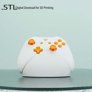 May include: A white and orange video game controller with a stand. The controller has a white base and orange buttons. The text ".STL Digital Download for 3D Printing" is visible at the top of the image.