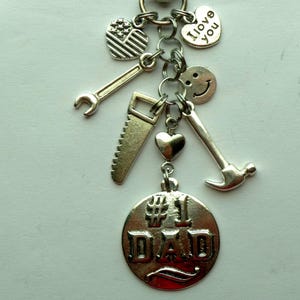 May include: A silver-toned keychain with various charms, including a wrench, saw, hammer, heart, smiley face, and a charm shaped like the American flag. The central charm reads "#1 DAD". The keychain is designed to be a gift.