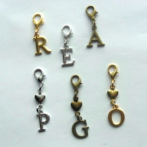 May include: A collection of six metal charms featuring letters and hearts attached to lobster claw clasps. The set includes gold-toned charms with letters R, A, and O, a silver-toned charm with letter E, a silver-toned charm with a heart and letter P, and a bronze-toned charm with a heart and letter G.