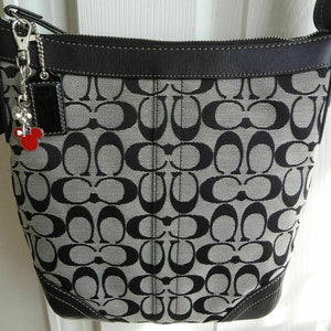 May include: A black and gray Coach shoulder bag with a repeating pattern. The bag has a black leather trim, a silver-tone strap, and a small red Mickey Mouse charm. The bag is made of a durable fabric and has a zipper closure.