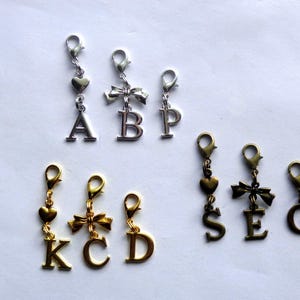 May include: A collection of metal letter charms with lobster claw clasps. The set includes silver-toned charms with letters A, B, and P, gold-toned charms with letters K, C, and D, and bronze-toned charms with letters S, E, and G, featuring heart and bow accents.