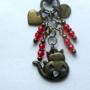 May include: A bronze-toned metal keychain charm featuring a cat-shaped pendant with heart cutouts, a solid heart charm, and a round disc stamped with the word LOVE. The design includes two dangling strands of faceted red beads and bronze spacers.