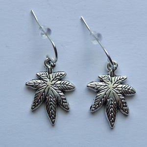 May include: A pair of silver-colored earrings. Each earring features a detailed, leaf-shaped charm suspended from a silver-tone ear wire. The charms have intricate veining details, and the earrings are set against a light background.