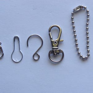 May include: An assortment of silver-colored metal findings, including a lobster clasp, a safety pin style clasp, a curved hook, a swivel clasp, and a ball chain. These findings are commonly used in jewelry making and crafting projects.