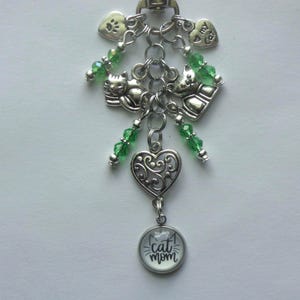 May include: A silver-toned keychain featuring a central chain with dangling charms including two cat figures, a filigree heart, and a circular pendant with the text cat mom. The design is accented with small green faceted beads and heart charms.