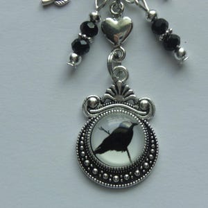 May include: A silver charm with a black bird silhouette in a glass dome, surrounded by silver beads. The charm is attached to a silver chain with black beads and a silver heart.