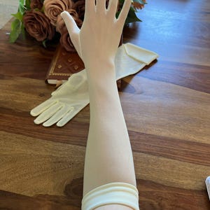 Könnte beinhalten: Ein Paar durchsichtige, elfenbeinfarbene Handschuhe. Einer der Handschuhe wird an einem Arm getragen und zeigt den zarten Stoff und die lange Länge. Ein passendes Paar Handschuhe und ein Rosenstrauß sind im Hintergrund. Klassisches Accessoire.