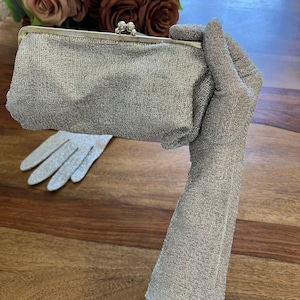 May include: A silver metallic clutch bag with a kiss-lock clasp closure, paired with a matching pair of long, shimmering silver metallic gloves. The fabric of both the clutch and the gloves features a fine, textured, glittery knit construction.