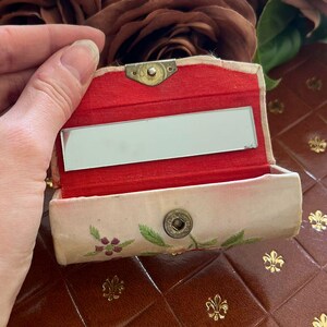 May include: A small, vintage, open compact mirror with a red interior and a floral design. The compact is held in front of a brown leather-bound journal and a bouquet of brown and tan roses.