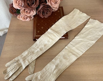 May include: A pair of long, white leather gloves. The gloves are laying on a wooden surface with a bouquet of brown and pink roses and a leather-bound book in the background.