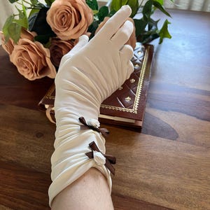 Op de afbeelding: Crèmekleurige handschoenen tot de elleboog met bruine satijnen strikken en kleine witte roosjes. De handschoenen zijn bij de pols gerimpeld en gemaakt van een zacht, mogelijk satijnachtig materiaal. Op de achtergrond een boeket perzikrozen en een leren boek.