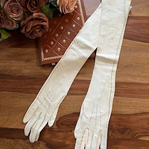 May include: Long, cream-coloured leather gloves with fine stitching, displayed on a wooden surface. Beside them is a brown leather-bound book with gold detailing and a bouquet of brown roses.