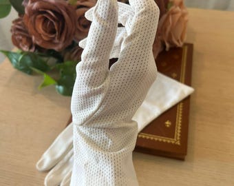 May include: A pair of white gloves with a perforated design. The gloves are held up in front of a bouquet of brown roses and a leather-bound book. The gloves have a classic, elegant style.