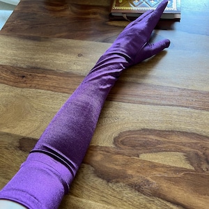 Peut inclure: Un long gant de style opéra en tissu violet vibrant et scintillant. Le gant dépasse le coude et présente une finition lisse, semblable à du satin, avec un léger éclat métallique qui capte la lumière. La matière est ajustée au bras.