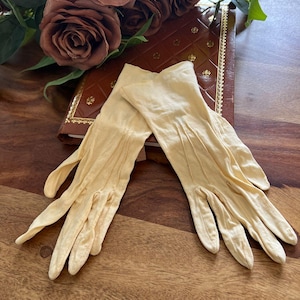 Può includere: Un paio di guanti vintage color crema con cuciture dettagliate. I guanti sono appoggiati su una superficie di legno accanto a un libro rilegato in pelle marrone e un mazzo di rose marroni.