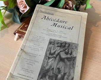 May include: A vintage book titled "Abécédaire Musical" by Maurice Chevais. The book cover features a black and white illustration of a group of children singing. The book is a first-year music textbook for students.