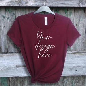 Op de afbeelding: Een bordeauxrood T-shirt met een ronde hals, hangend aan een zwarte hanger tegen een verweerde houten achtergrond. De tekst "Your design here" is in cursieve letters op de voorkant van het T-shirt geschreven.