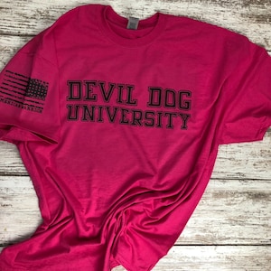 Op de afbeelding: Een roze T-shirt met de tekst "DEVIL DOG UNIVERSITY" in zwarte letters. De linkermouw heeft een verweerde Amerikaanse vlag met de tekst "AMERICAN WARRIOR" eronder.